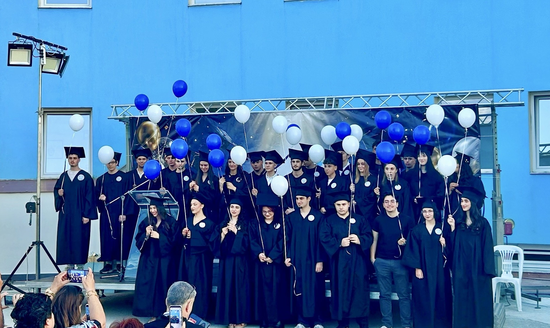 Group of graduates in black gowns and mortarboards on a stage, holding blue and white balloons, as audience captures photos.