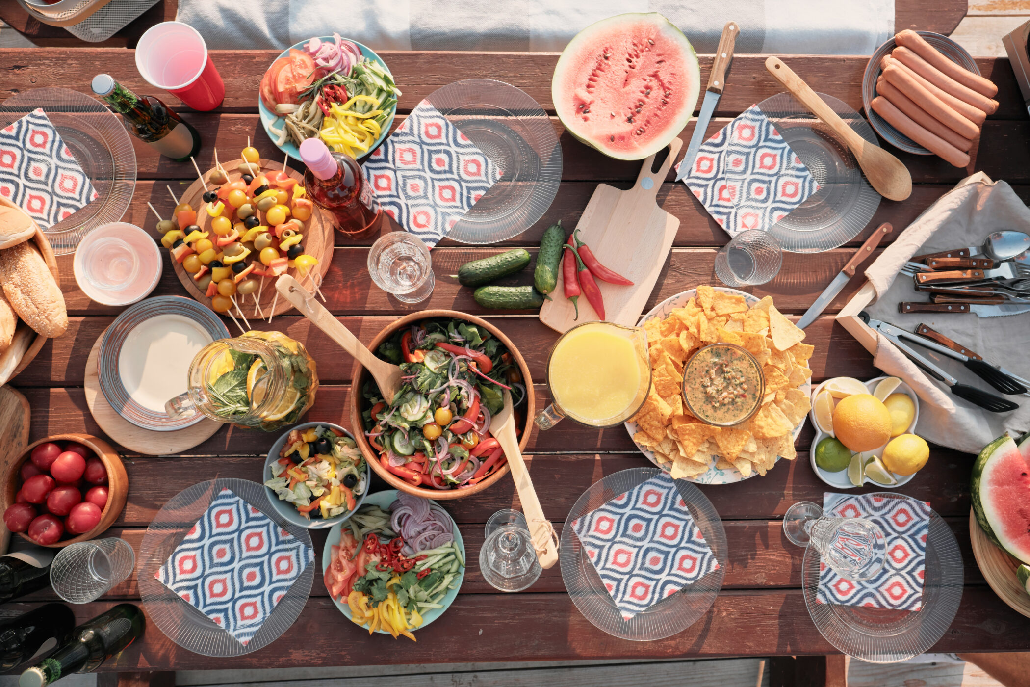 Overhead view of a wooden picnic table loaded with plates of salad, fruit, sausages, chips, watermelon, and drinks.