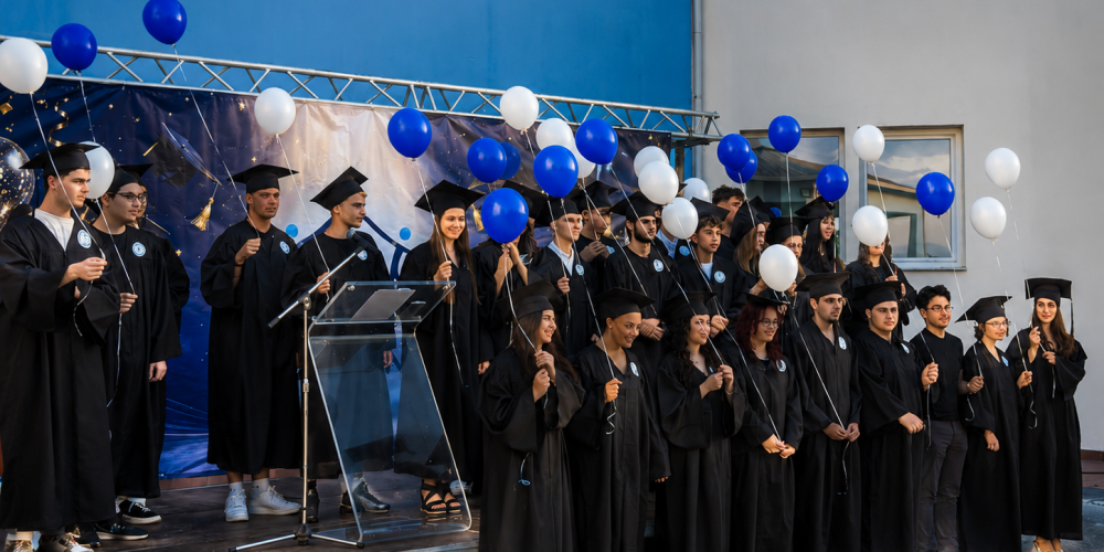 Graduates in black robes and mortarboards on a stage outdoors, holding blue and white balloons during a graduation ceremony.