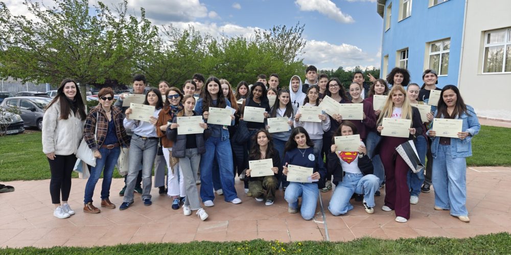 Group of smiling students holding certificates posing outside a blue school building on a sunny day with trees behind them.