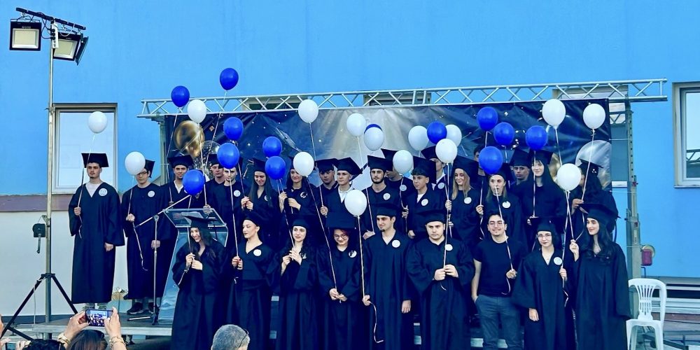 Group of graduates in black gowns and mortarboards on a stage, holding blue and white balloons, as audience captures photos.