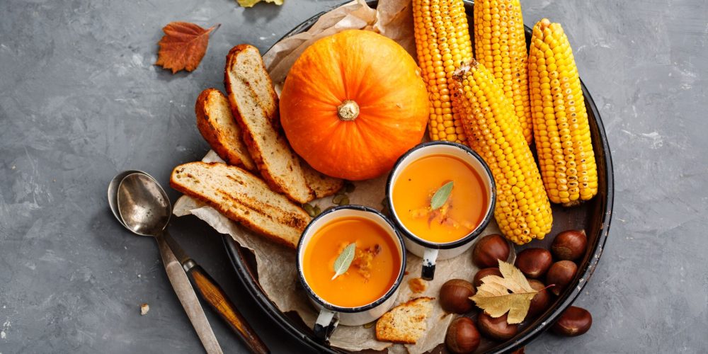 Autumnal spread with pumpkin, corn on the cob, toasted bread and bowls of orange soup on a dark tray.