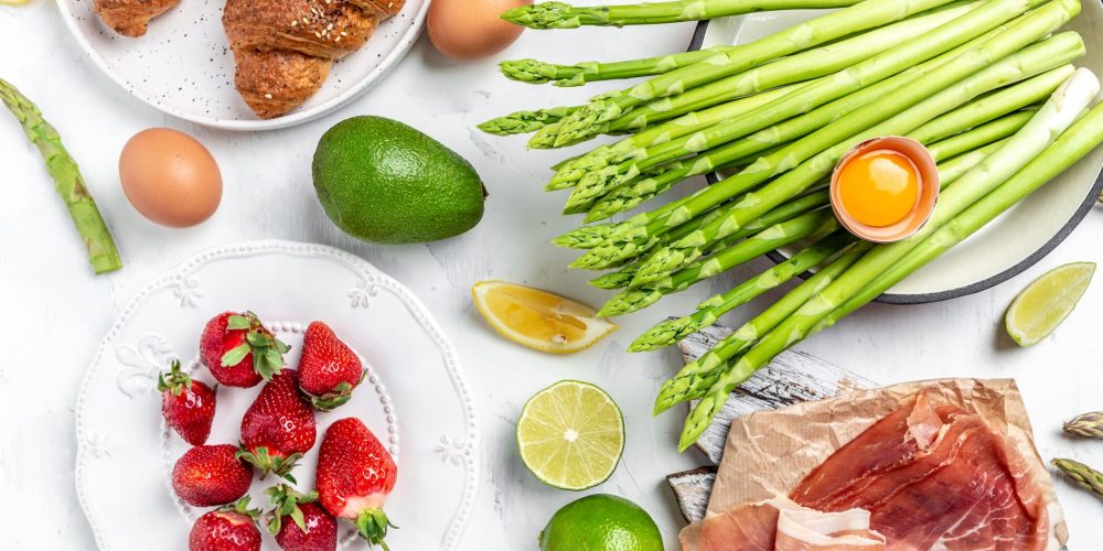 Top-down view of a breakfast spread with croissants, asparagus, eggs, avocado, berries, citrus wedges, and sliced ham on a white surface