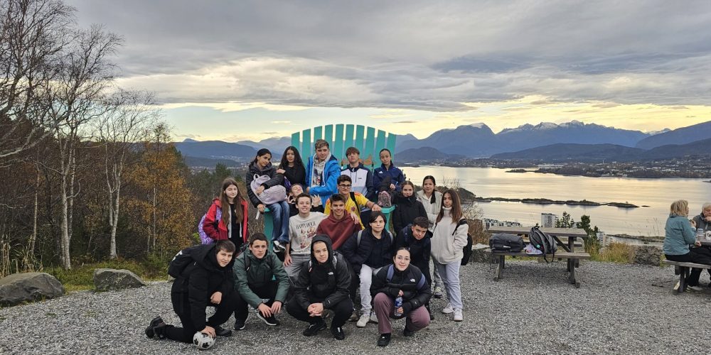 Group of students posing for a photo at a hilltop overlook with a lake and snowy mountains in the distance under a cloudy sky, many wearing backpacks and jackets.
