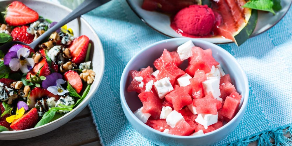 Bowl of watermelon and feta cheese cubes beside a colorful leafy salad with strawberries and edible flowers on a blue placemat.