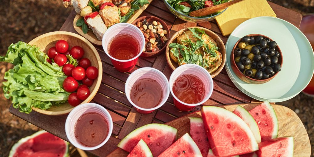Outdoor picnic spread on a wooden table with watermelon slices, fresh greens, cherry tomatoes, olives, salad bowls, skewers, nuts, and red drinks in plastic cups in a sunny outdoor setting.