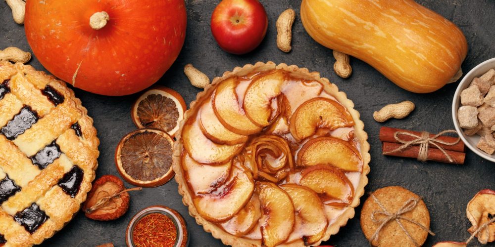 Assorted fall pastries and fruits on a dark surface: a large orange pumpkin, a red apple, a pale orange squash, a pear-topped tart, dried orange slices, peanut shells, cinnamon sticks, and a bowl of sugar cubes in the corner.
